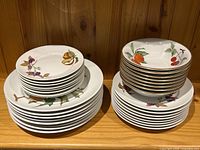 Four stacks: dinner plates, salad plates, dessert plates, bowls, all with fruit motifs and gold rims