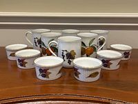 Six Royal Worcester Evesham porcelain coffee mugs and six ramekin dishes arranged in rows, showing front fruit design patterns.