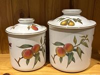 Pair of white porcelain storage jars with lids featuring hand-painted fruit designs (peach and pear) with gold trim, photographed against wood panel background.