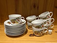 Eight porcelain cups stacked beside eight matching saucers with fruit pattern and small candle holder on wooden shelf