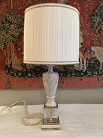 Full view of the lamp showing the urn-style marble body with pleated white shade and ivory fabric-wrapped cord against a tapestry background.