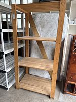Photo of wooden IKEA storage shelf with 4 wooden tiers showing front angle view with adjacent shelving units and mattress in background.