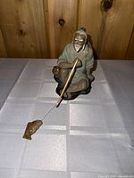 Front and side view of the pottery figurine depicting an old man fishing, showing detailed facial features and fishing pole with fish.