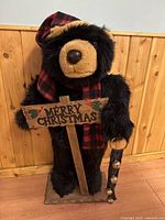 Full view of the black plush Christmas bear standing on a wooden base, wearing a plaid red and black scarf and stocking cap, holding a wooden 'Merry Christmas' sign and a strap of jingle bells.
