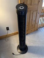 Full height view of the black Seville Classics tower fan showing the vertical vents, circular base, and power cord on carpeted floor near wooden door.