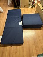 Photo showing one navy blue trifold mattress pad fully unfolded and another folded into a compact trifold block on hardwood floor.