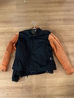 Full front view of black body riding coat with orange sleeves hanging on hanger on wooden floor.