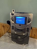 Front view of Karaoke Vision STVG535 karaoke machine showing built-in screen illuminated in blue with microphone plugged in and control buttons on front panel.