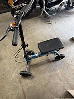 Photo showing the full KneeRover knee scooter from above on concrete floor, blue frame, black padded seat, four wheels visible.