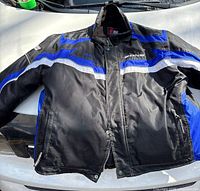 Front view of men's Zone motorcycle jacket in black with blue and white details, sized XL, laid on vehicle hood showing front zipper and pocket details