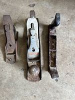Three assorted hand planers on concrete floor, including a Stanley branded block plane with a wooden knob, adjacent to two other metal planers, one with a black handle.