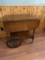 Wooden vintage style tea cart with left drop leaf folded down, side view showing wooden spoke wheels and caster wheels.