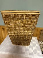 Front and side view of rectangular wicker laundry hamper on table showing woven pattern and lid with wooden rim.