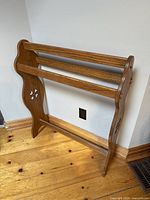 Side angled view of the wooden quilt rack showing three horizontal bars and decorative cutout sides, standing on wood floor.