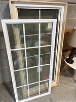 Photo showing two new white framed windows leaning against a concrete wall, one window laid in front of the other showing full rectangular shape with grid pattern in glass panes.