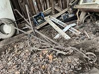 Wide view of antique metal plow resting on dirt floor amidst leaves and debris in a storage area showing overall frame, blade, and wheel.