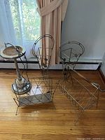 Four vintage metal items including a metal ashtray stand with marble base, 2 umbrella holders, and a magazine holder on hardwood floor near window.