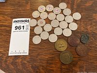 Top view of 32 assorted coins and tokens including Canadian commemorative quarters with pink ribbons and other varied tokens on a wood surface with MaxSold bidding card visible