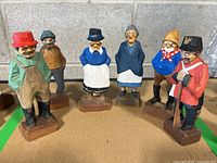 Six folk art wood carvings displayed on a table against a gray wall, showing all individual figures in the set.