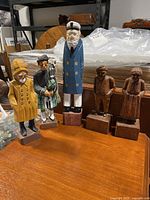 Five folk art wood carvings displayed on a wooden table, showing the variety of figures including a ship's captain, Scottish man, and three unpainted figures.