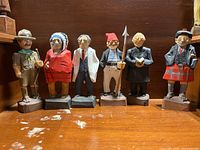 Six folk art wood carvings featuring various characters including a scout, Native American chief, man in suit, pirate, priest, and man in Scottish clothing, arranged side by side.