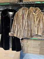 Three fur coats hanging on a metal rack: one long black faux fur coat with collar and lining, two short mink fur jackets in brown shades with visible buttons