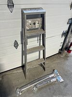 Full view of aluminum step ladder standing upright with two packaged bathroom mobility assist grab bars on the ground.