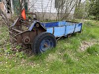 Manure spreader trailer parked on grass with blue sides, single axle with one visible tire, some rust and faded paint, orange reflective slow-moving vehicle sign attached to rear.