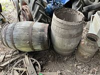 Photo showing two large wooden barrels lying on the ground and one vintage metal milk can placed beside them. Surrounding farming equipment and tires are visible in background.