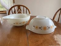 Two white milk glass Pyrex mixing bowls on a wooden table, showing the orange and brown geometric floral pattern.