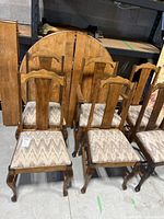 Photo showing round wooden table top leaning against a storage wall with 4 rectangular leaves beside it, and six dining chairs in front with medium wood finish
