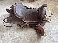 Full view of worn brown leather horse saddle showing seat, pommel, cantle, and attached stirrups on floor.