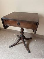 Side view of small mahogany drop leaf pedestal table showing drawer with metal lion pull and metal feet on tripod base.