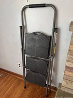 Folded Cosco 3-step ladder showing plastic steps and metal frame with a padded handle.