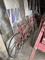 Full side view of the red metal tandem bicycle frame leaning against wood and other materials in a storage area.