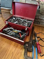 Red metal toolbox with lid open showing sockets, wrenches, pliers, and various hand tools in drawer and on top.