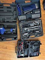 Overview of all tools in their cases on wooden floor, showing rotary tool, ratchet sets, and nailer