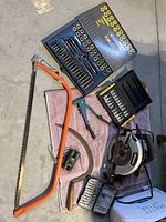 Tools including tap and die set, circular saw, hand scythe, riveter, and binoculars displayed on a floor.