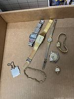 Lot of assorted vintage wristwatches, a gold-tone linked bracelet, and a silver-tone shield-shaped prayer keychain in a cardboard box, showing varying sizes and styles of watches with visible wear and age.
