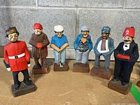 Six carved and painted wood figurines by Tom Hannah, displayed in a row showing different characters.