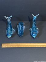 Top and side views of 3 blue swirl and clear glass dolphin figurines with a ruler for scale, each approximately 6 inches long.