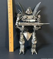 Front view of welded steel robot art figure with ruler indicating height about 10 inches, showing detailed mechanical design and long rod held in hands.