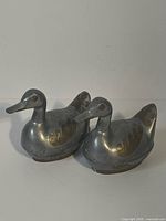 Front and side view of two metal duck figurines made of brass and pewter with decorative wing accents.