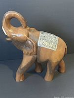 Front-side angled view showing the elephant's curved raised trunk, wooden carved body with textured details, and silver tone metal plate on the back.
