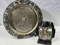 Metal plate featuring Aztec calendar center and abalone shell border next to black ceramic mask with inlaid shells on face