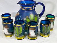Photo of six blue glasses arranged around the blue glass pitcher, each piece showing unique hand painted cactus and Mexican figure designs.