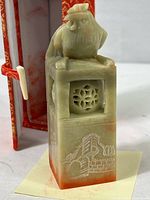 Front view of the greenish soapstone stamp with a rooster atop and carved designs including the Great Wall on the front. The stamp is standing upright next to the open red and gold fabric-covered box.