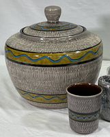 Punch bowl with textured gray body, banded with yellow and blue wavy design, with lid and knob top next to a single cup showing matching design.