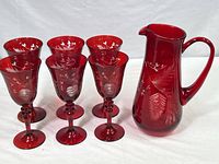 Front view of one ruby red glass pitcher with etched pinecone and evergreen branch patterns, alongside six matching goblets, all in good condition.