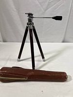 Full view of the tripod standing with its legs extended, showing brown wooden legs with chrome caps and the attached carrying case lying next to it.